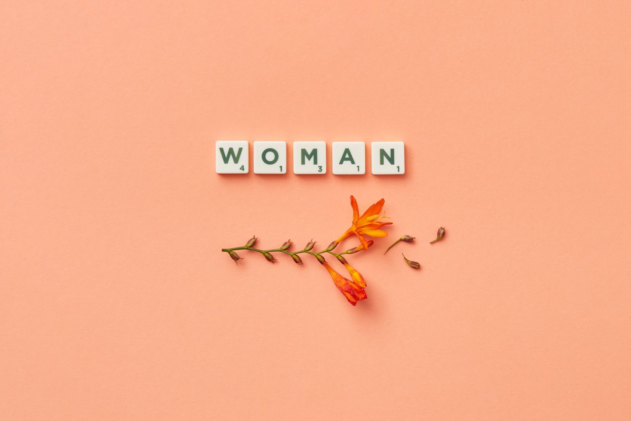 A minimalist composition featuring Scrabble tiles spelling woman with delicate orange flowers on a peach background.